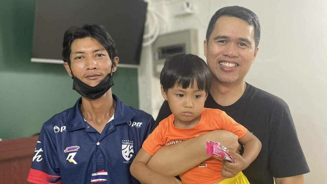 A man named Khun Nai standing in a welcoming gesture, smiling at Pastor Mark and his wife Jenny. The image captures a moment of gratitude and faith, symbolizing a transformative journey from illness to salvation through the power of God's grace.