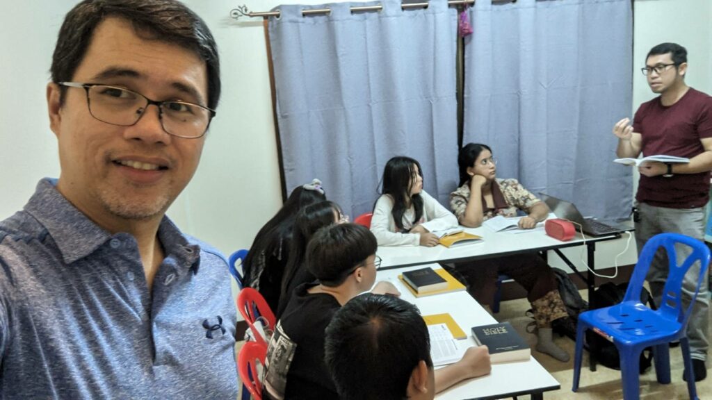 A man named Khun Nai standing in a welcoming gesture, smiling at Pastor Mark and his wife Jenny. The image captures a moment of gratitude and faith, symbolizing a transformative journey from illness to salvation through the power of God's grace.