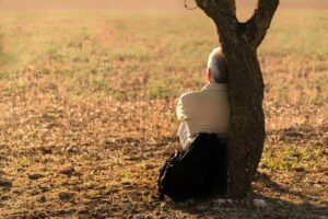 image of a man sitting in an open field leaning on the tree experiencing the life of loneliness.