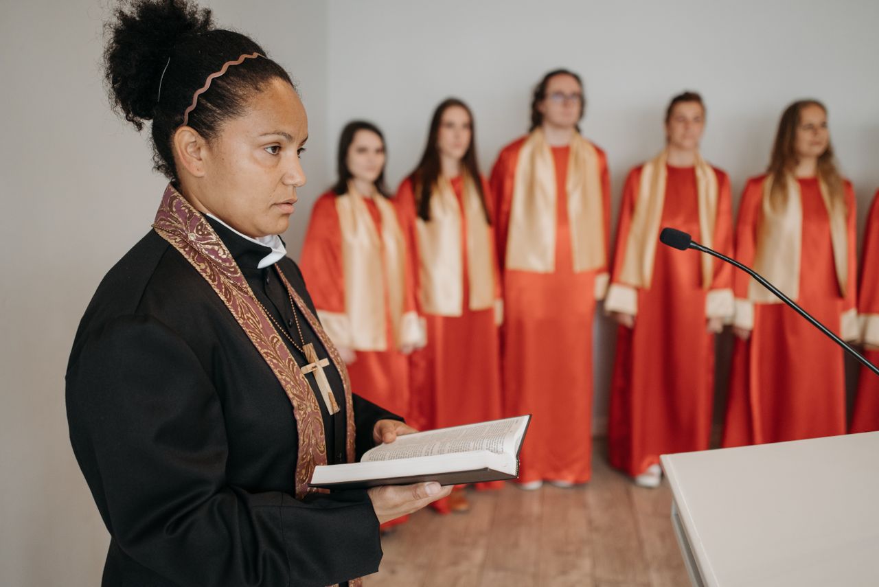 A female pastor holding a Bible leads a choir in red robes during a church service.